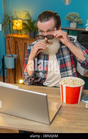 Kaukasischer älterer Mann Freiberufler macht Pause von der Arbeit mit 3D-Brille, isst Popcorn und schaut Film auf Laptop. Lachender Großvater, der am Schreibtisch sitzt und in der Arbeitspause Snacks trinkt Stockfoto