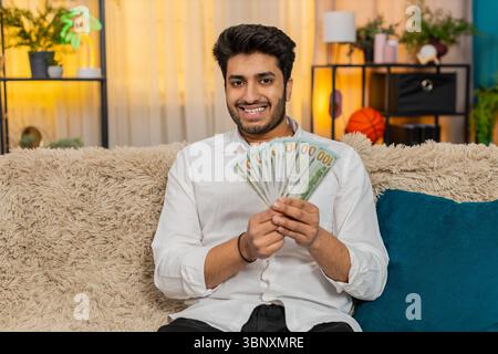 Indischer Mann zu Hause, der Bargeld zählt, sein Budget sorgfältig überlegt und zukünftige Ausgaben plant. Ein Araber sitzt auf dem Sofa und konzentriert sich auf Geldmanagement und durchdachte Finanzentscheidungen Stockfoto