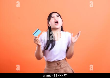 Asiatische Frau frustriert nachdem sie ihre Kreditkarte gesehen hat, ist sie mit ihren Finanzen gestresst und sieht verzweifelt aus, isoliert auf orangem Hintergrund Stockfoto