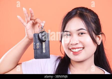 Eine junge asiatische Frau lächelt, während sie einen Autoschlüssel und eine Koffer in der Hand hält. Steht für den Besitz eines neuen Fahrzeugs, isoliert auf orangefarbenem Hintergrund. Stockfoto