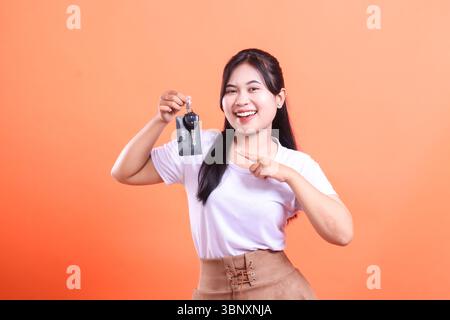 Glückliche junge Frau, die Autoschlüssel hochhält und mit dem Finger zeigt, stolz lächelt und in die Kamera blickt. Isoliert auf orangefarbenem Hintergrund Stockfoto