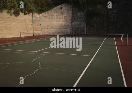 Die flache Oberfläche des Tennisplatzes zeigt Risse, weiße Linien und ein durchhängendes Netz mit Lampenpfosten Stockfoto