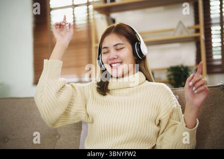 Junge asiatische Frau mit Kopfhörern, die sich zu Hause entspannen. Musik hören. Sitzen auf einem gemütlichen Sofa mit Kopfhörern Stockfoto