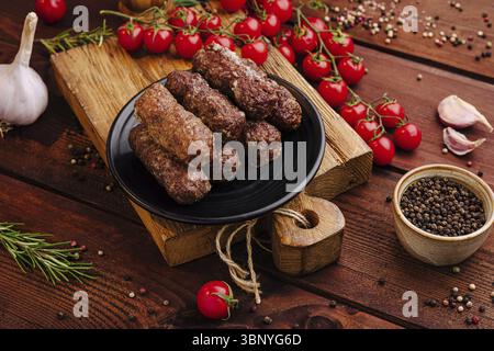 Food still-Life, saftige Fleischspieße sind auf einem schwarzen Teller angeordnet, umgeben von lebhaften Kirschtomaten, Knoblauchzehen und verschiedenen Gewürzen auf einem Rusti Stockfoto