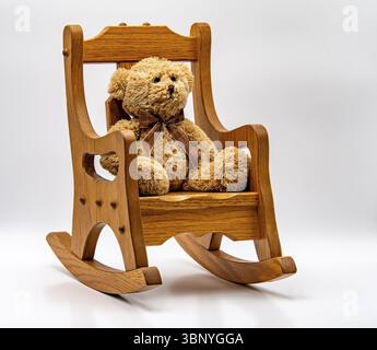 Ein weicher Teddybär mit braunem Band sitzt bequem auf einem Holzschaukelstuhl in einem sauberen, minimalistischen Innenraum. Das Ambiente insgesamt ist Stockfoto