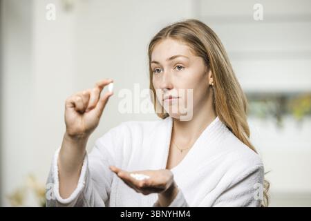 Junge, blonde Frau in einem weißen Bademantel mit einer Handvoll Medikamente, skeptischer Blick im Spiegel Stockfoto