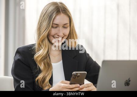 Junge, blonde Frau in schwarzem Blazer sitzt mit Handy und Laptop am Tisch, lächelt Stockfoto