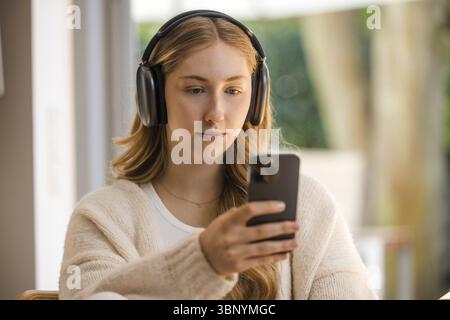 Junge, blonde Frau in weiß sitzt entspannt am Tisch mit Kopfhörern und Handy und konzentriert sich Stockfoto