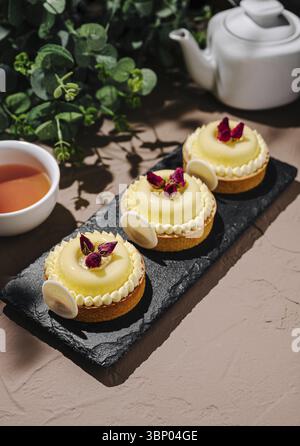 Food still Life, drei wunderschön gefertigte Zitronentörtchen sitzen auf einem schwarzen Schieferbrett, jede mit Rosenblättern und weißer Schokolade belegt. Eine Tasse Kräutertee Stockfoto