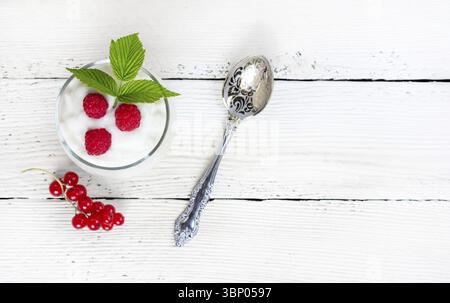 Ein Glas mit Milchjoghurt, Reifen Himbeeren, grünem Blatt und roter Johannisbeere auf einem weißen Holztisch mit Kopierbereich von oben, selektiver Fokus Stockfoto