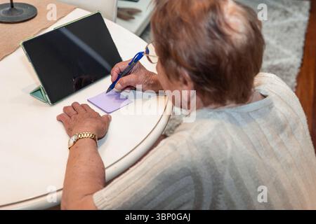 Eine ältere Frau schreibt Notizen auf ein kleines Papier an einem Tisch mit einem Tablett in der Nähe. Die Szene spiegelt einen gemütlichen und organisierten Lebensstil in ihrem Zuhause wider, Highli Stockfoto