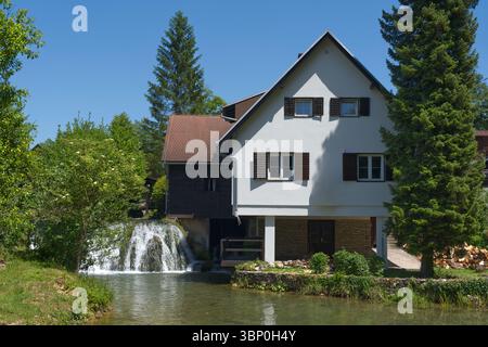 Ein bezauberndes weißes Haus mit braunen Fensterläden steht neben einem fließenden Wasserfall und einem ruhigen Fluss Stockfoto
