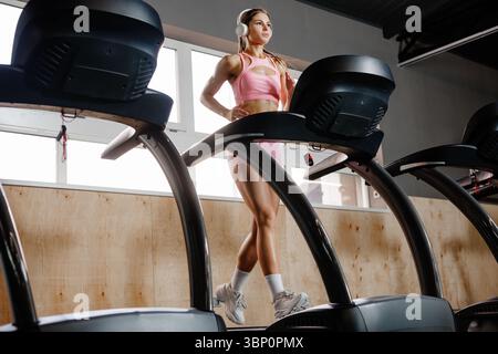 Athletische Frau, die auf dem Laufband im Fitnessstudio läuft, trägt Kopfhörer Cardio Fitness Trainer gesunde Lebensweise Workout Motivation drinnen Stockfoto