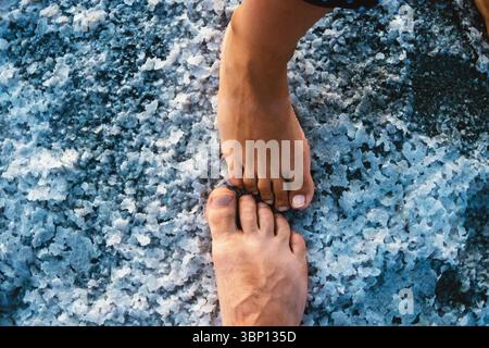 Zwei Beine, weiblich und männlich, berühren die zerrissene Oberfläche eines ausgetrockneten Salzsees. Das Bild konzentriert sich auf die Interaktion zwischen den Füßen und dem Unique Tex Stockfoto