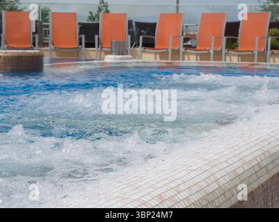 Ein Blick aus der Nähe auf einen sprudelnden Whirlpool mit weißen und braunen Mosaikfliesen, mit verschwommenen orangefarbenen Lounge-Sesseln im Hintergrund unter klarem Hintergrund Stockfoto