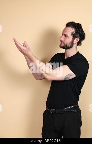 Ein junger Mann in einem schwarzen T-Shirt und einer Hose, im Seitenprofil gesehen, blickt mit beiden Händen nach oben und offenen Handflächen nach oben, als ob man Wiegemöglichkeiten hätte. Stockfoto