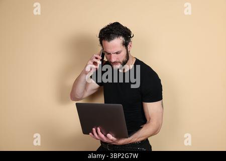Ein fokussierter Mann kann mehrere Aufgaben ausführen, indem er mit seinem Smartphone spricht, während er gleichzeitig arbeitet und seinen Laptop-Bildschirm vor einem schlichten beigen Hintergrund betrachtet. Stockfoto