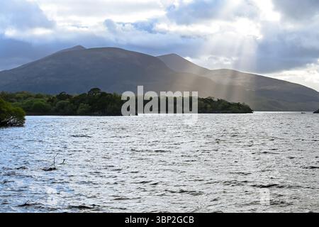 Die Sonne scheint durch Wolken über einem See mit Bergen Stockfoto