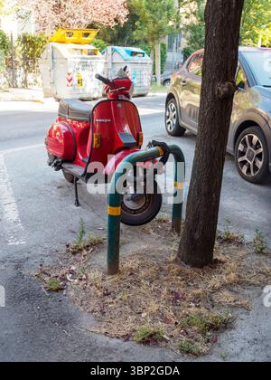 Bologna, Italien 3. Juli 2025 Red Vespa PX mit Kettenschloss um das Rad, das neben einem Baumstamm und einigen Mülltonnen in einem Wohngebiet, urb, geparkt ist Stockfoto