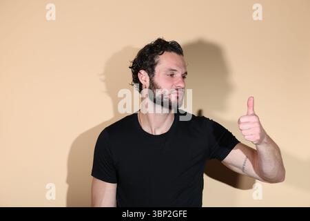 Ein junger bärtiger Mann mit einem schwarzen T-Shirt steht vor einem schlichten Studio-Hintergrund, gibt eine klare Geste der Zustimmung und sieht vertrauenswürdig aus Stockfoto