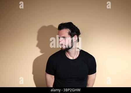 Ein bärtiger Mann in einem schwarzen T-Shirt wird in einem Studio festgehalten und blickt nachdenklich über seine Schulter nach links, weg von der Kamera, gegen einen Stockfoto