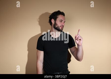 Ein bärtiger Mann in einem schwarzen T-Shirt schaut nachdenklich nach oben, während er seinen Zeigefinger nach oben zeigt, was auf eine Idee oder einen Schlüsselpunkt hindeutet, Agi Stockfoto