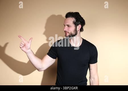 Ein selbstbewusster bärtiger Mann in einem schwarzen T-Shirt macht mit seiner Hand eine Fingerpistolen-Geste, während er nach rechts blickt und eine spielerische oder selbstbewusste Attit vermittelt Stockfoto