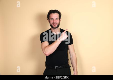 Ein freundlicher junger Mann in einem schwarzen T-Shirt und einer Hose lächelt die Kamera an, während er mit dem Zeigefinger nach oben und zur Seite zeigt, als ob er etwas anzeigt Stockfoto