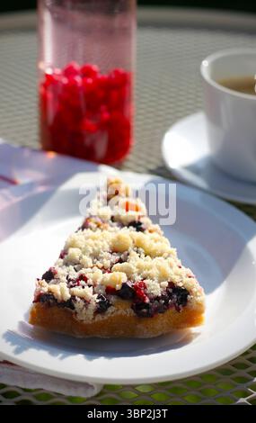 Hausgemachte rote und schwarze Johannisbeercrumb-Torte auf einem weißen Teller, mit Beeren-Limonade und Tasse Kaffee im Hintergrund, Sommer-Dessert-Konzept Stockfoto