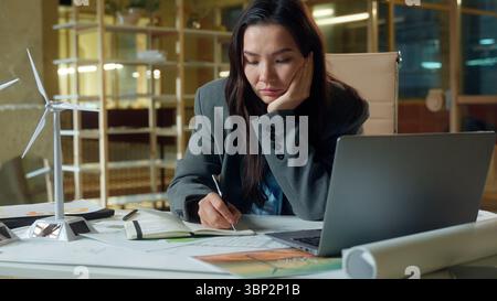 Traurige unglückliche asiatische Geschäftsfrau Architektin Ingenieur Geschäftsfrau schreibt Notizen im Büro Windmühle Elektrokraftprojekt nachdenkliche nachdenkliche Verärgerung Stockfoto