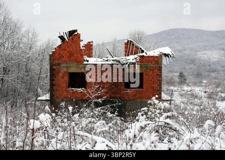 Ein teilweise eingestürztes Haus aus rotem Backstein steht verlassenen, schneebedeckten Feldern, umgeben von blattlosen Bäumen und fernen Hügeln unter grauem Winterhimmel Stockfoto