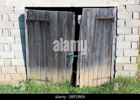 Zwei alte, dunkle Holztüren mit blauem Seilverschluss sind in einer rauen Betonblockwand angebracht und zeigen Anzeichen von Verschleiß, Sonnenlicht und bewachsenem Gras Stockfoto