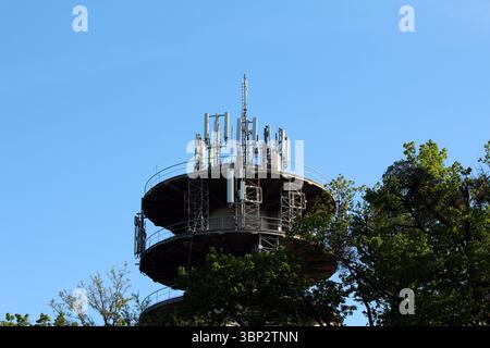 Über grünen Bäumen erhebt sich ein kreisförmiger Telekommunikationsturm, der mit zahlreichen Antennen und Sendern für die Mobilfunk- und Funkkommunikation ausgestattet ist Stockfoto