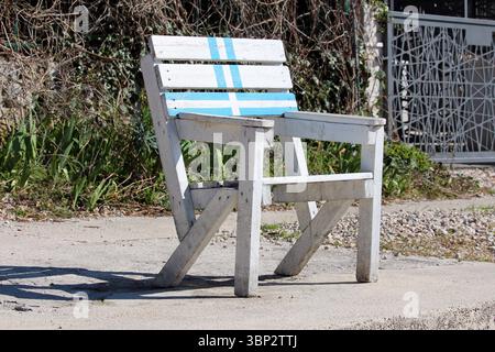 Ein rustikaler weißer Holzstuhl mit kräftigen blauen Streifen steht allein auf einem sonnendurchfluteten Betonweg, dessen alte Farbe und robuste Bauweise ein Gefühl von Outdoor wecken Stockfoto