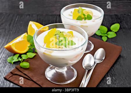 Dessert aus Joghurt mit Orange, Banane und Kiwi in zwei Glasschalen, Früchten, Löffeln und Minze auf einer braunen Serviette auf Holzbrett Hintergrund Stockfoto