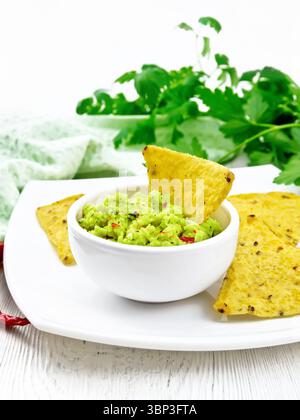 Guacamolensauce in einer Schüssel mit Maischips auf Teller, heißen Pfefferschoten, Handtuch und Petersilie auf dem Hintergrund eines hellen Holzbretts Stockfoto