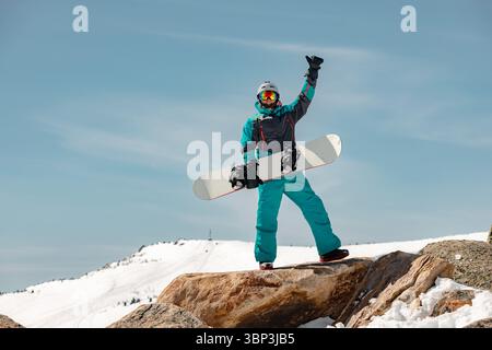 Ein männlicher Snowboarder steht auf einem Felsen mit erhobenem Arm vor dem Hintergrund einer verschneiten Piste hoch in den Bergen Stockfoto