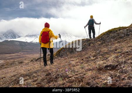 Zwei weibliche Trekkingerinnen mit Rucksäcken und Trekkingstöcken stehen an einem Berghang und bewundern die schneebedeckten Gipfel des hohen Altai-Gebirges am bewölkten Herbsttag. Konzept für aktive Ferien Stockfoto