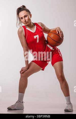Selbstbewusste junge Frau in roter Uniform posiert mit Basketball und symbolisiert Sportlichkeit, Teamwork und sportliche Motivation. Stockfoto