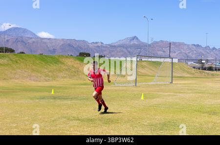 Männlicher Fußballspieler in roter Uniform sprintet auf dem Grasfeld und weicht gelben Kegeln durch ein Metalltor aus Stockfoto