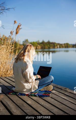 Frau, die am Laptop arbeitet, während sie auf einem Holzsteg in der Nähe des Wassers sitzt und die Freiheit genießt, fernzuarbeiten. Freiberufliche Tätigkeit Stockfoto