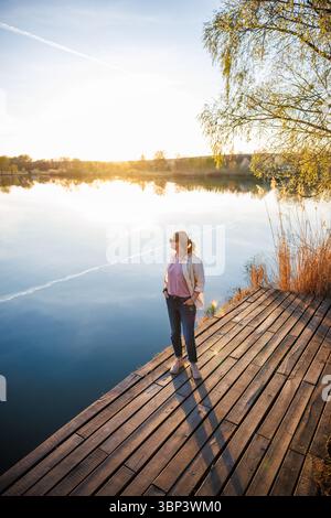 Frau, die bei Sonnenuntergang auf dem Pier am See steht. Genießen Sie die goldene Stunde und den ruhigen Moment in der Natur Stockfoto