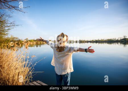 Sorglose Frau mit Hut und ausgestreckten Armen genießt die ruhige Landschaft am See. Freude an der Freiheit in der Natur Stockfoto