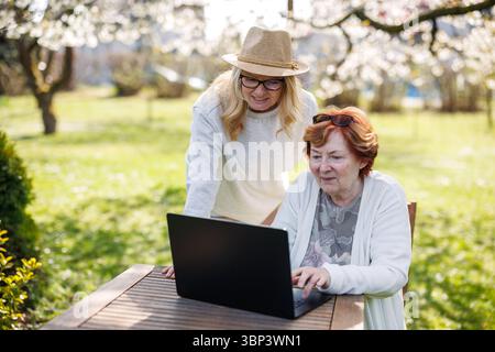 Glücklicher Moment, als eine Frau mit ihrer älteren Mutter lächelt, während sie draußen den Laptop-Bildschirm ansieht Stockfoto