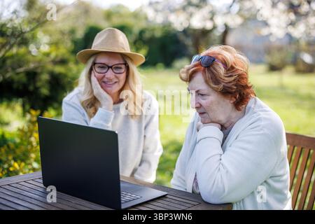 Glücklicher Moment, als eine lächelnde Frau mit ihrer älteren Mutter beim Anschauen des Laptops im Garten war Stockfoto