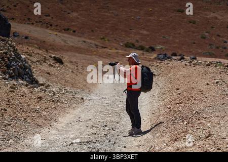 Mann wandert über felsiges vulkanisches Gelände mit Kamera in der Hand und Berglandschaft im Hintergrund Stockfoto