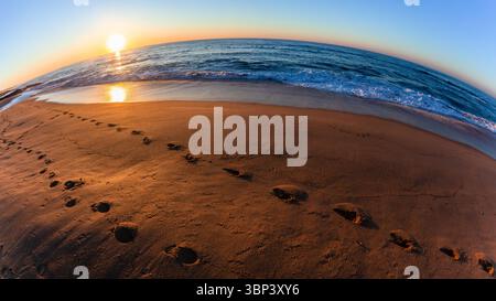 Das Wasser des morgens am Strand umrandet Fußabdrücke in Sand, während die Sonne über der gekrümmten Landschaft des Ozeans aufgeht. Stockfoto