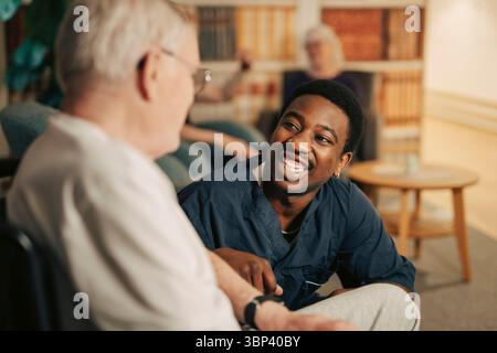 Lächelnde junge Krankenschwester, die mit einem älteren Mann im Rollstuhl im Altersheim kommuniziert Stockfoto