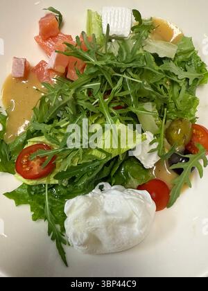 Das Foto zeigt einen frischen gemischten Salat, der auf einem weißen Teller serviert wird. Es enthält Rucola, Römersalat, Kirschtomaten, grün und schwarz Stockfoto