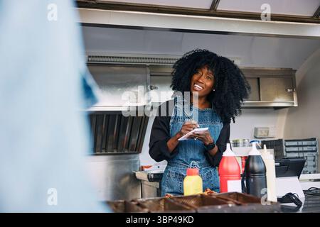 Eine weibliche Besitzerin mit lockigen Haaren schreibt die Bestellung des Kunden auf, während sie im Food Truck steht Stockfoto